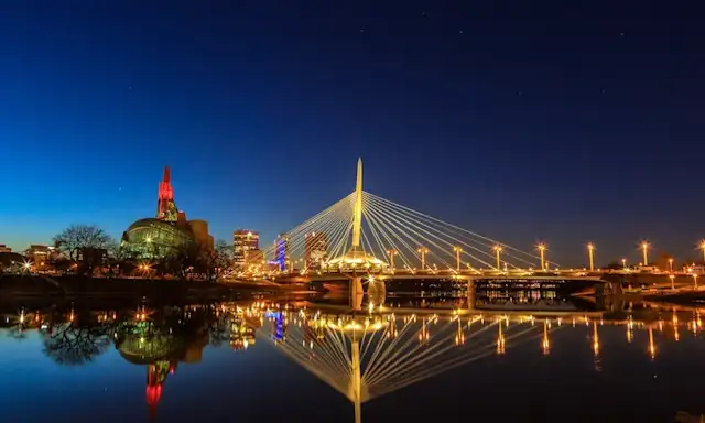 Night view of the Esplanade Riel pedestrian bridge in Winnipeg, Manitoba, illuminated with golden lights, reflecting in the river below.