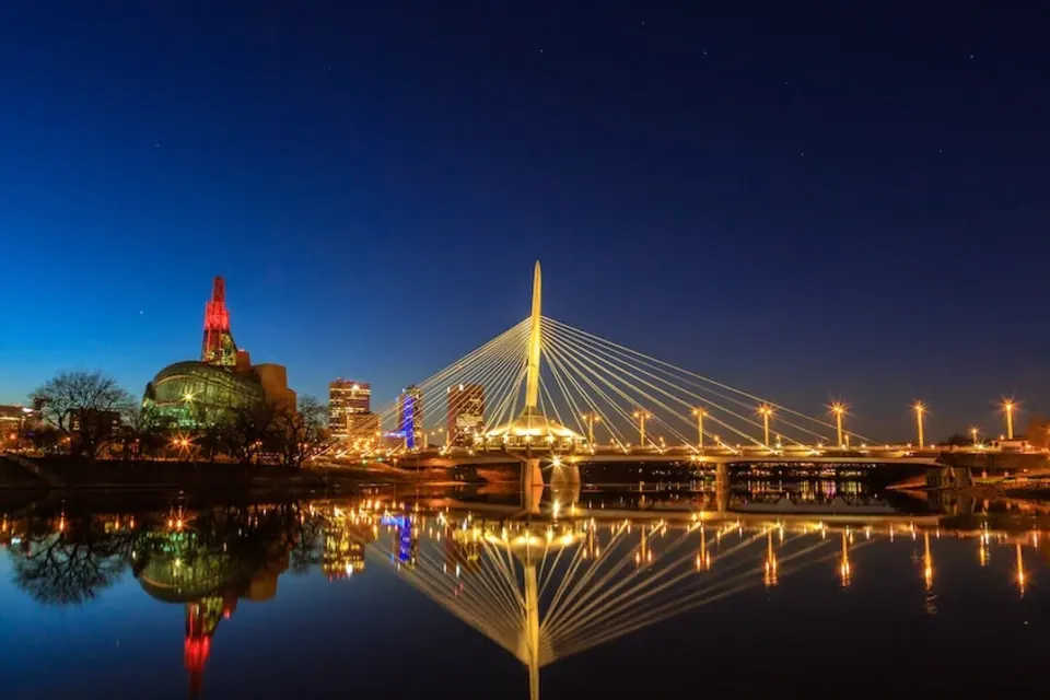 Night view of the Esplanade Riel pedestrian bridge in Winnipeg, Manitoba, illuminated with golden lights, reflecting in the river below.