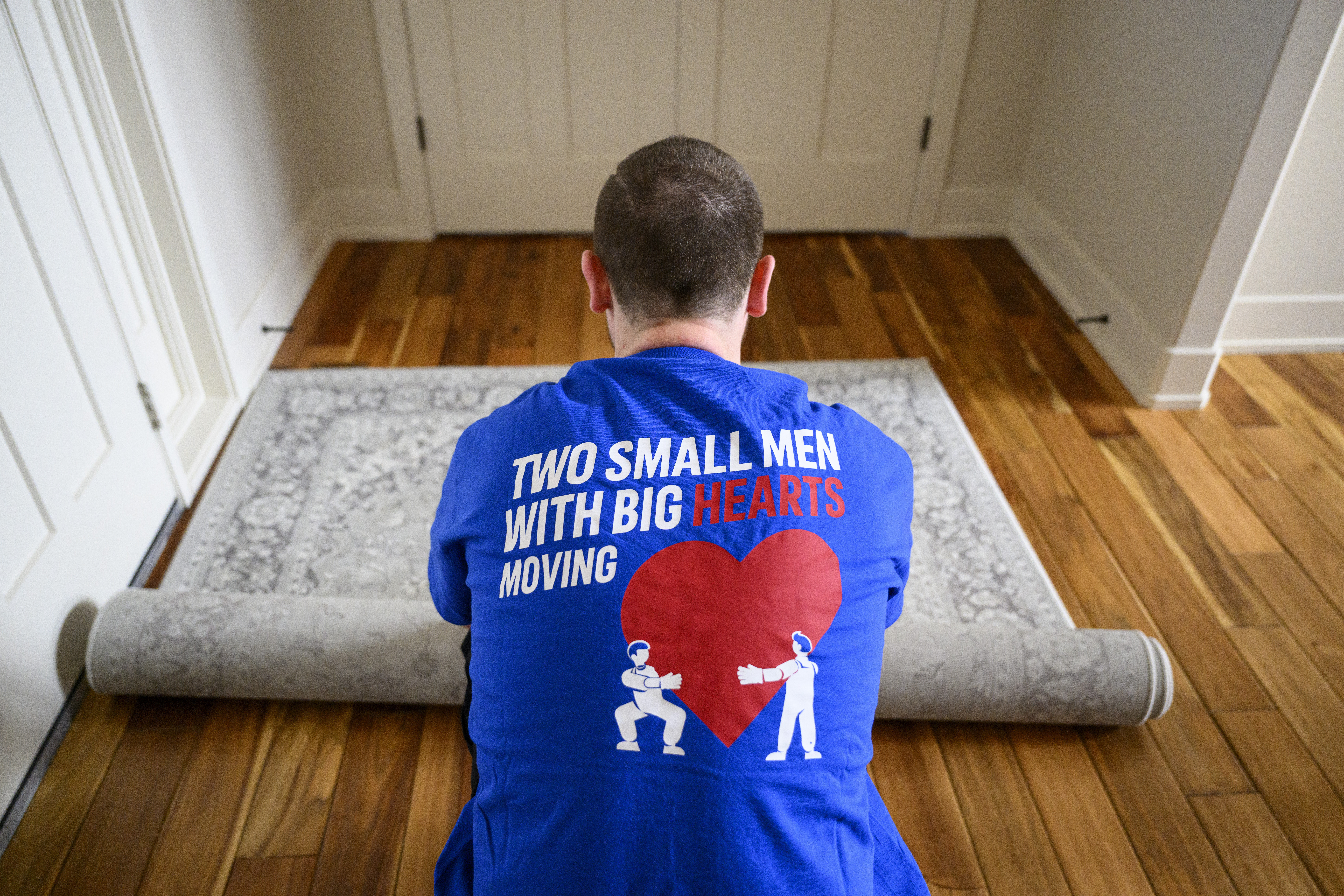Man in a Two Small Men with Big Hearts Moving shirt rolling up a rug on a wooden floor.