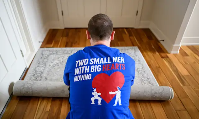 Man in a Two Small Men with Big Hearts Moving shirt rolling up a rug on a wooden floor.