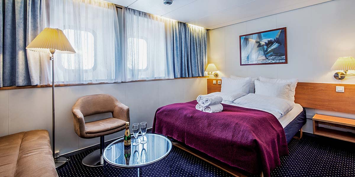 Wnętrze kabiny Commodore z dwuosobowym łóżkiem, lampą i fotelem, obok stolik z szampanem