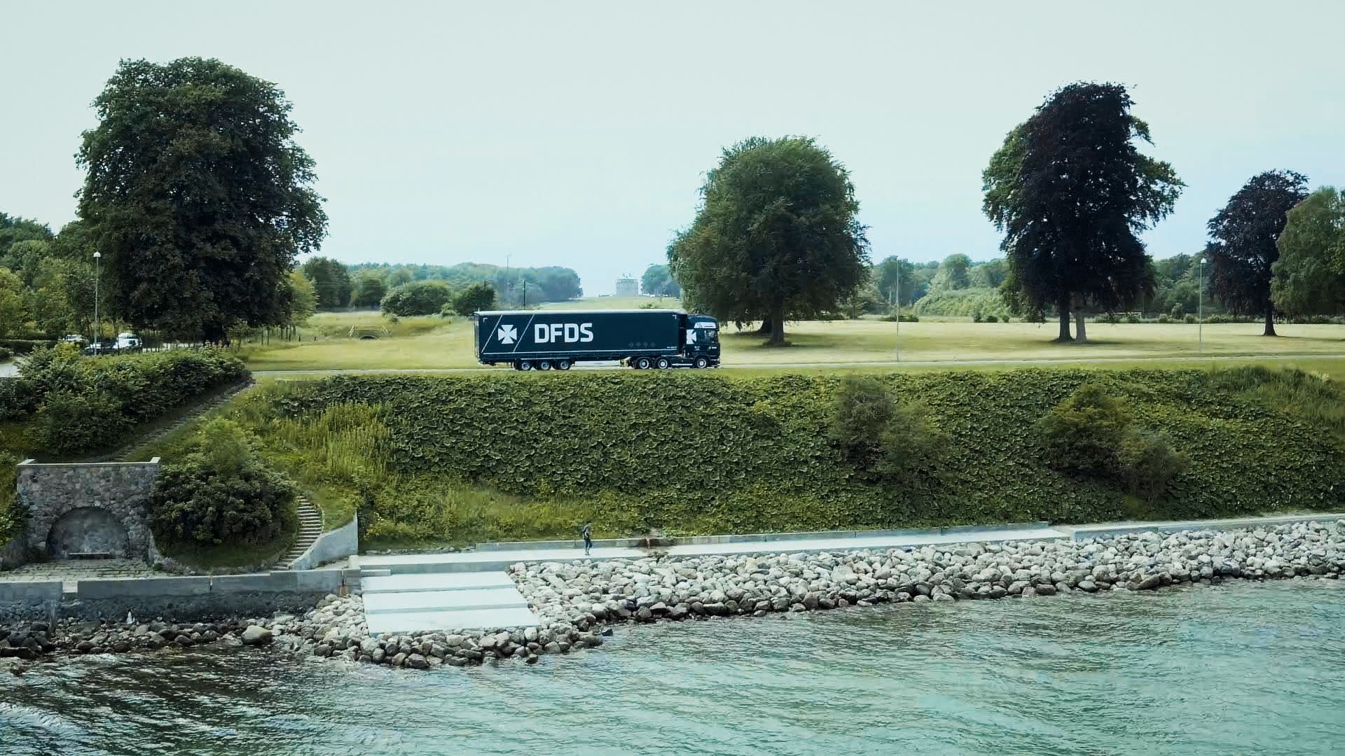 A DFDS Logistics truck driving though the countryside, surrounded by trees and a river shore DFDS-logistics-norway