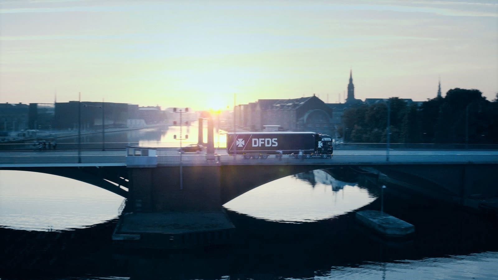 A blue DFDS Freight and Logistics truck drives over a bridge in front of a city at sunrise DFDS sunset