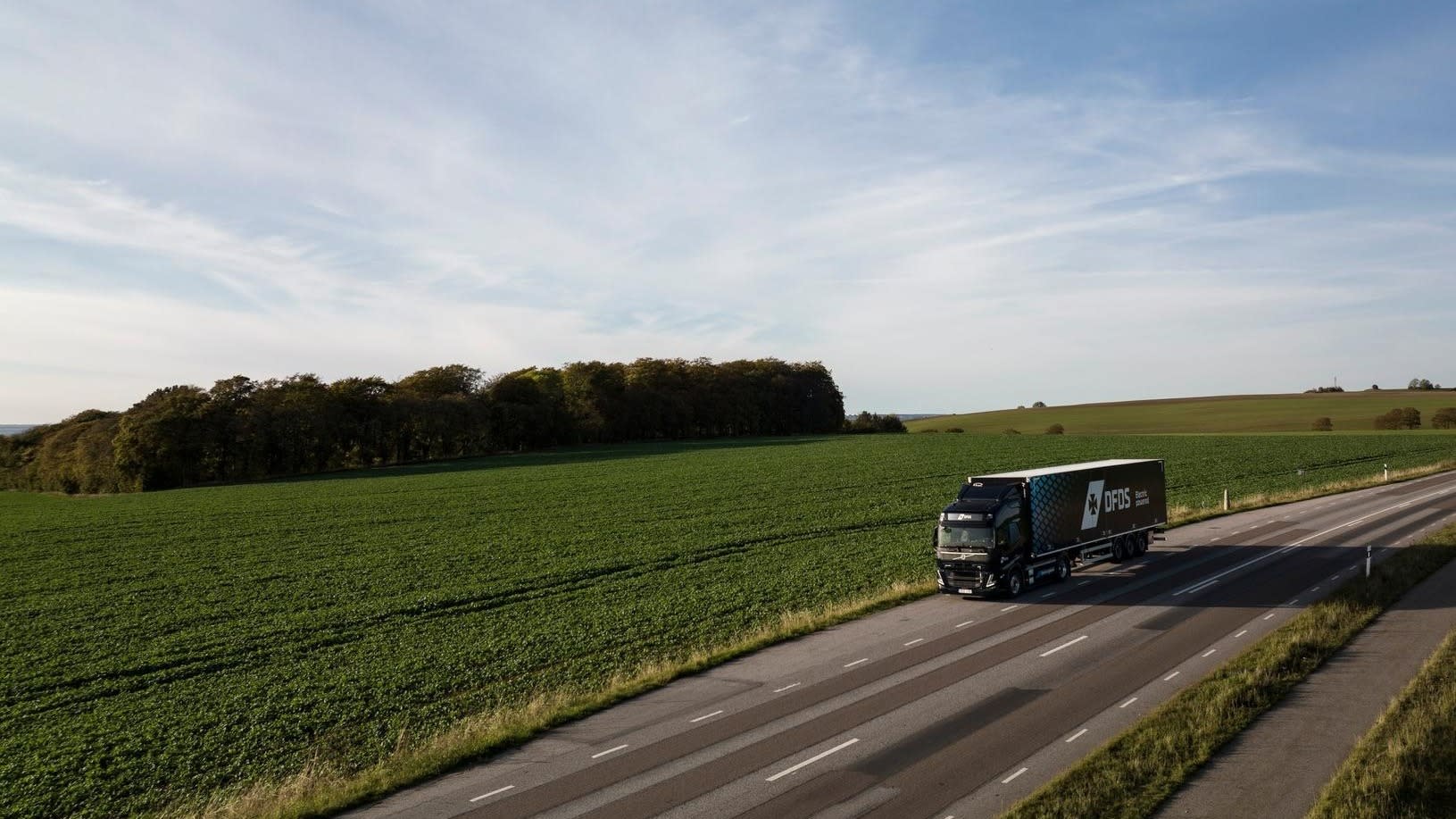 Purpose & Strategy page, About section, Pillar D Move to Green - electric DFDS truck in dark blue livery driving on the road, in a green scenery 86216-DJI 0031 1702066991366