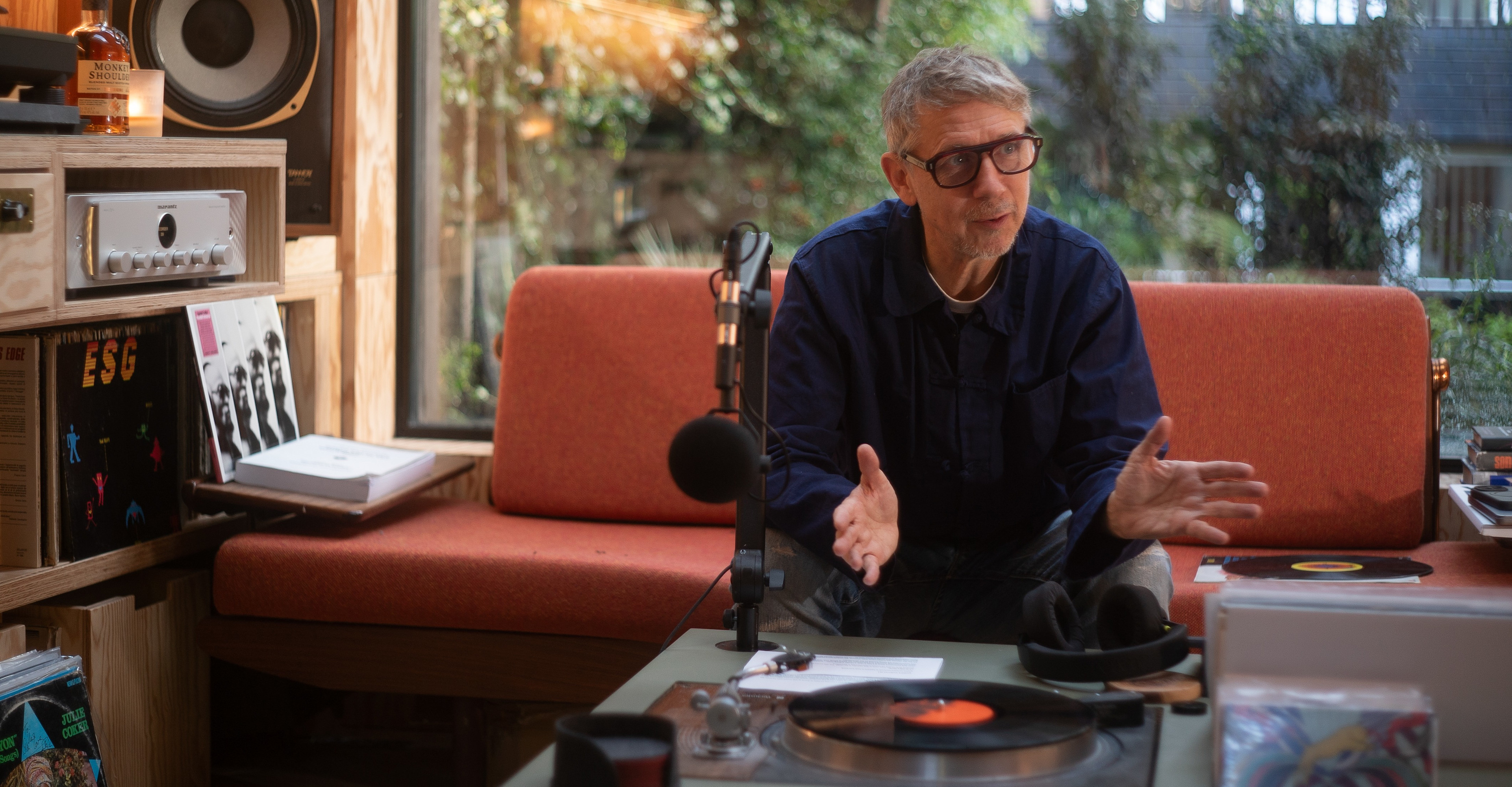 Gilles Peterson (a broadcaster and DJ from Worldwide FM radio) sitting on a sofa by a microphone, vinyl record shelf and vinyl record player