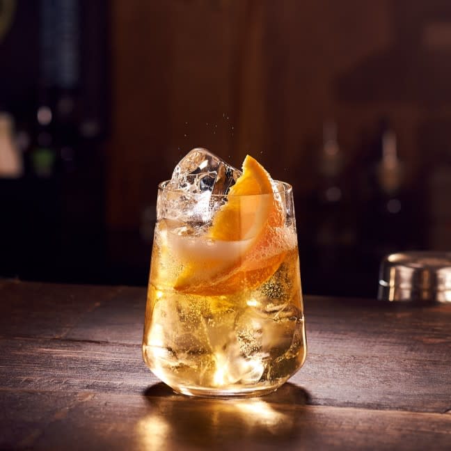 A Smokey Splash whisky and soda cocktail served with ice and an orange wedge sitting on a wooden bar