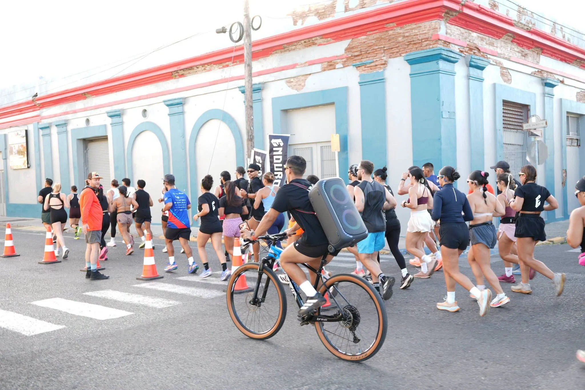 Corredores y ciclista pasan frente a edificio azul y blanco con molduras rojas durante carrera en la calle.
Foto vía Facebook: Ciudad de Ponce