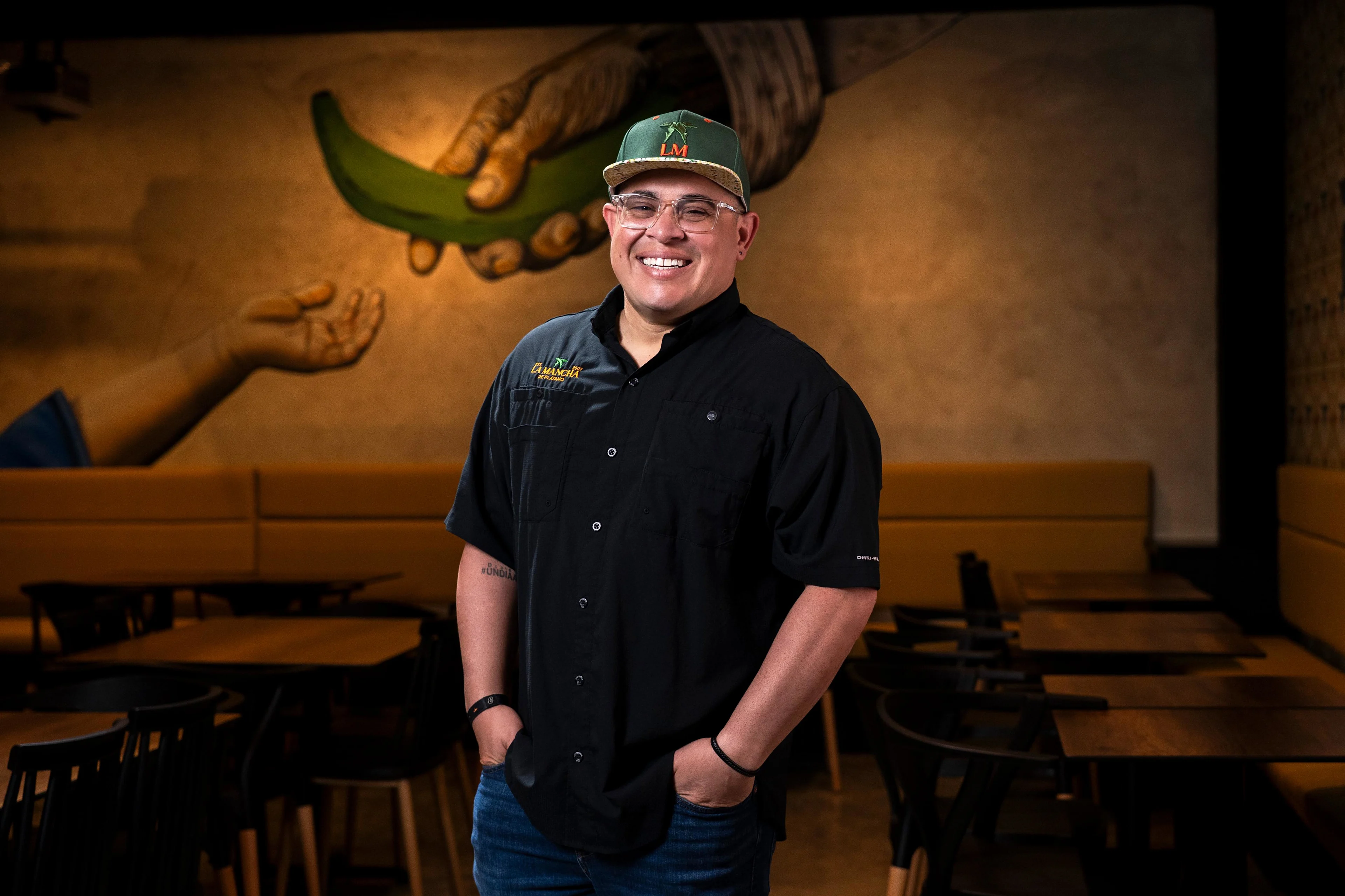 Chef sonriente con gorra verde y camisa negra frente a mural de plátano verde y manos en restaurante La Mancha de Plátano.
Emanuel Ocasio, fundador y chef ejecutivo de La Mancha de Plátano