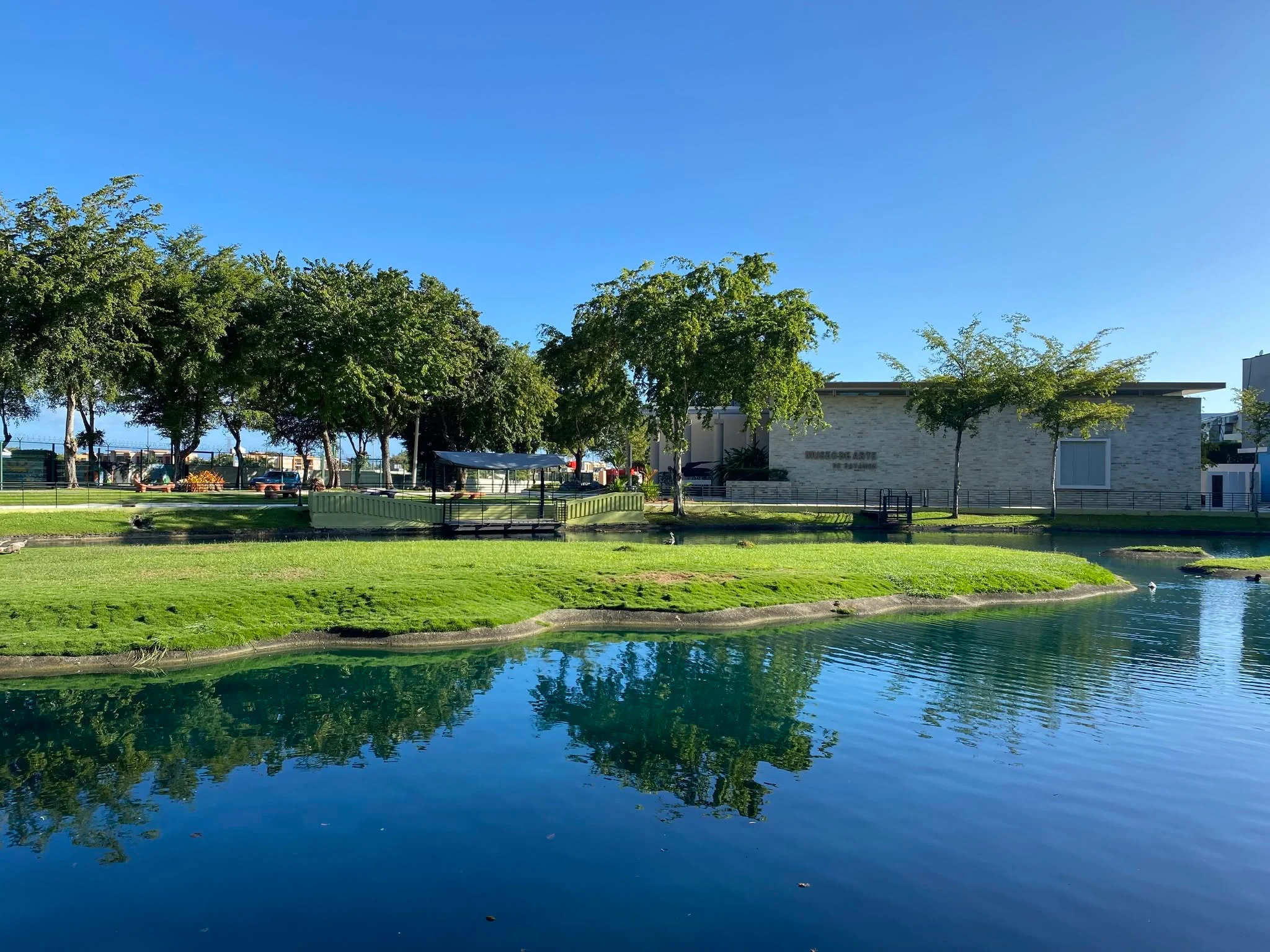 Laguna con césped verde, árboles y edificio de museo de arte bajo cielo azul despejado en día soleado.
Foto: Facebook: Museo de Arte de Bayamón