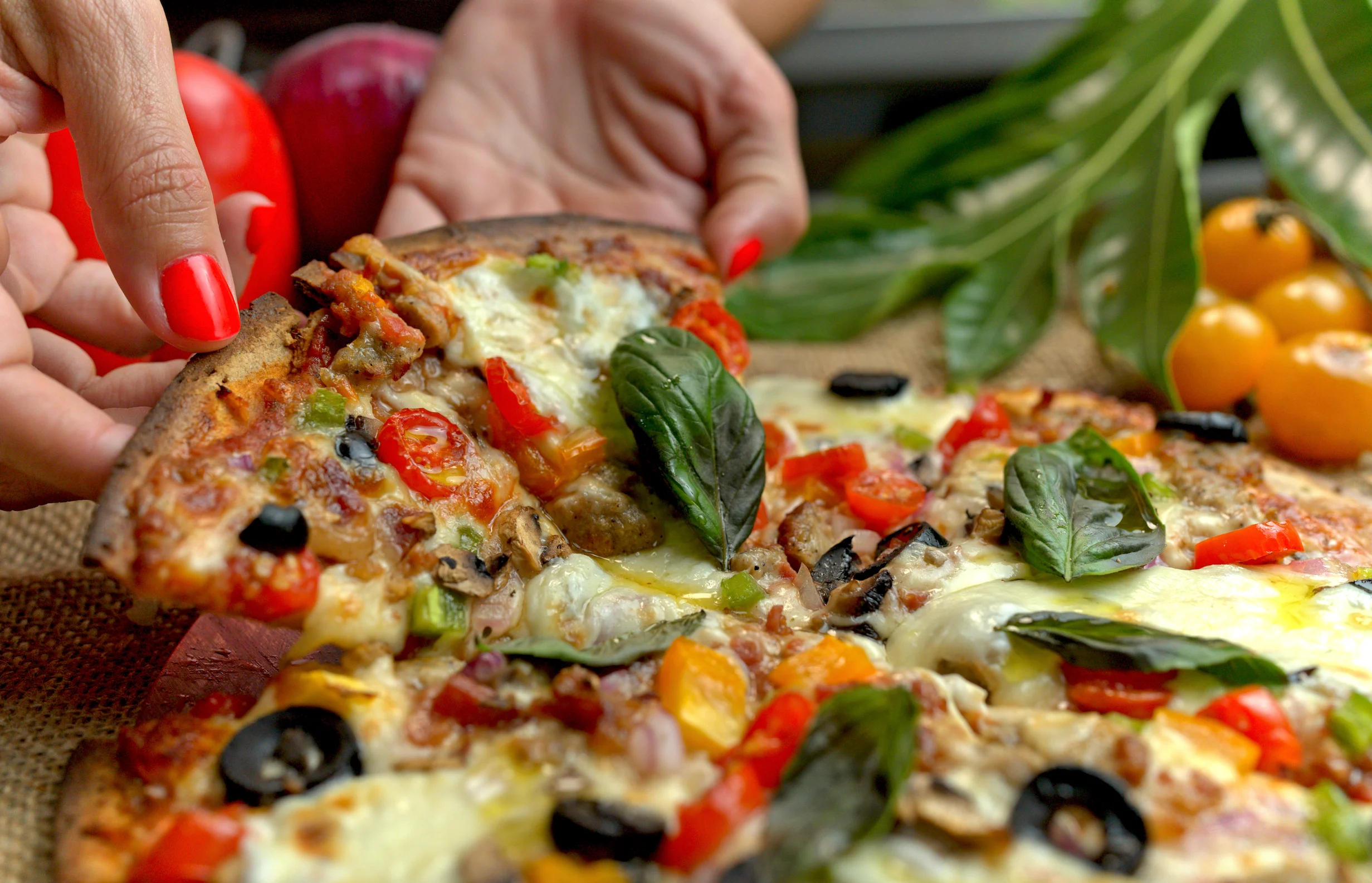 Mano con uñas rojas levanta una tajada de pizza con vegetales, aceitunas negras, queso derretido y albahaca fresca

Foto: Amasar