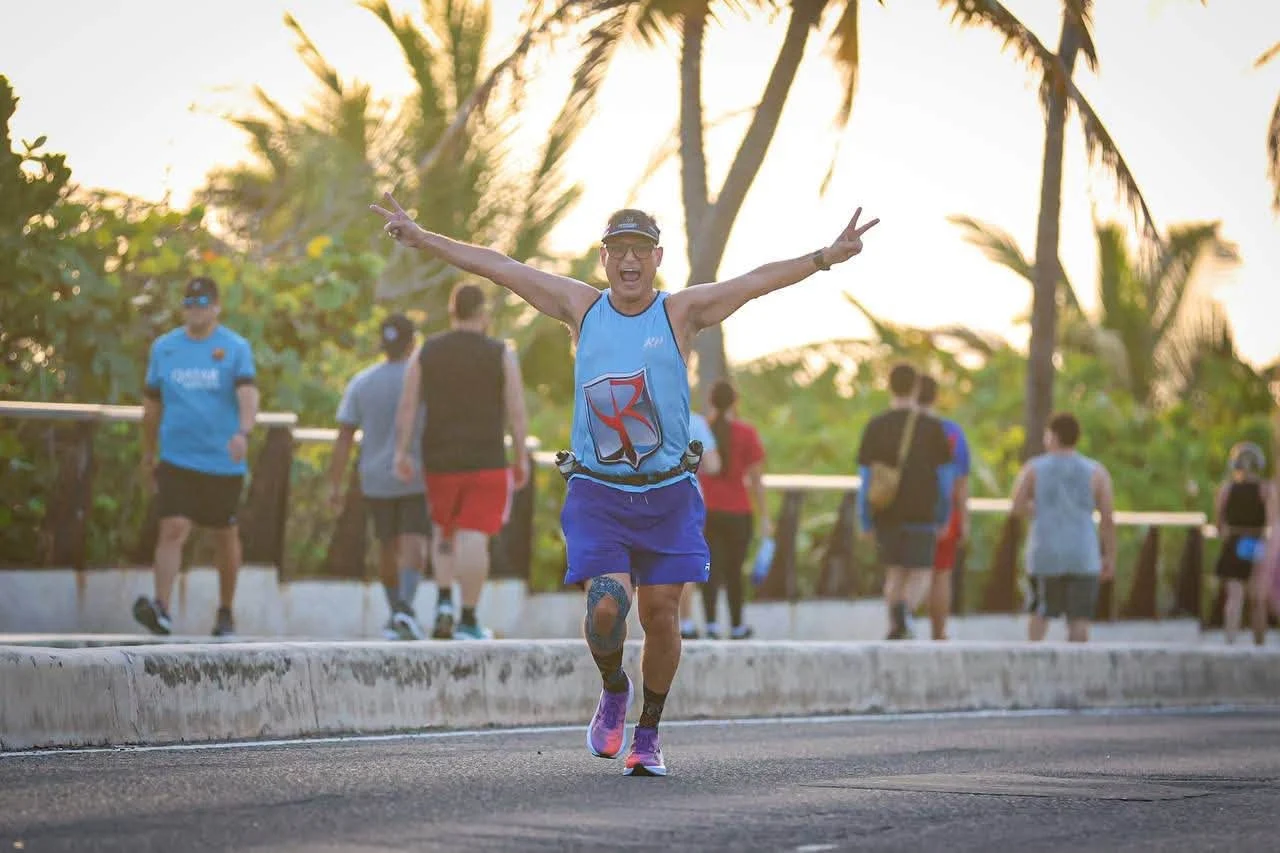 Corredor con camiseta azul y gorra celebra con los brazos abiertos en carrera al aire libre con palmeras de fondo
Foto vía Facebook: San Juan, Ciudad Capital