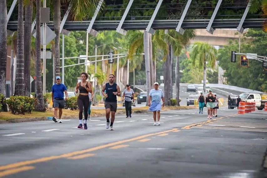 Grupo de personas corriendo y caminando en una avenida con palmeras y estructura metálica sobre la vía en San Juan
Foto Vía Facebook: San Juan, Ciudad Capital
