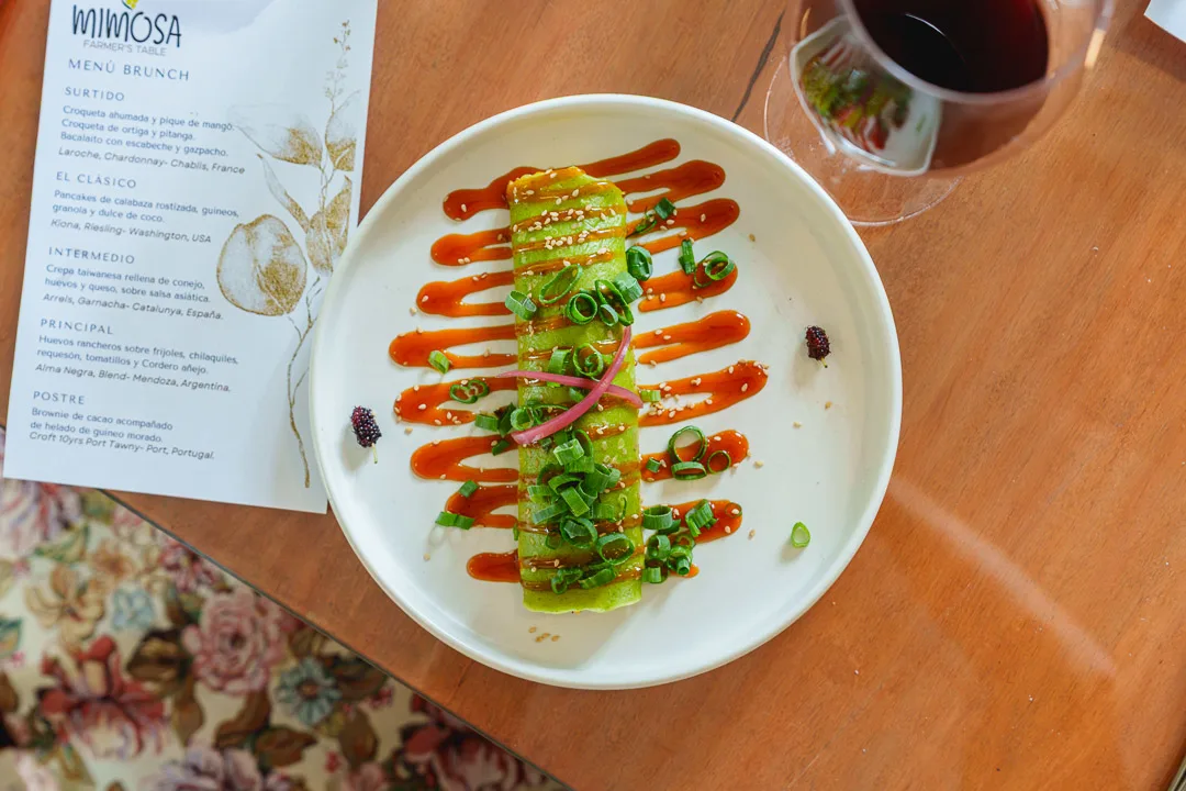 Plato de brunch con tostadas de aguacate decoradas con salsa roja, cebollín y encurtidos junto al menú de Mimosa