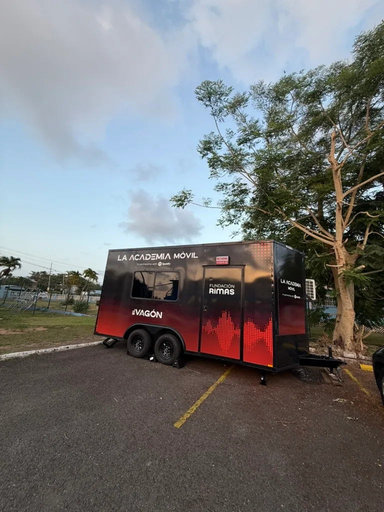 Tráiler negro y rojo de La Academia Móvil de Fundación Rimas estacionado en un área abierta con árboles al fondo.