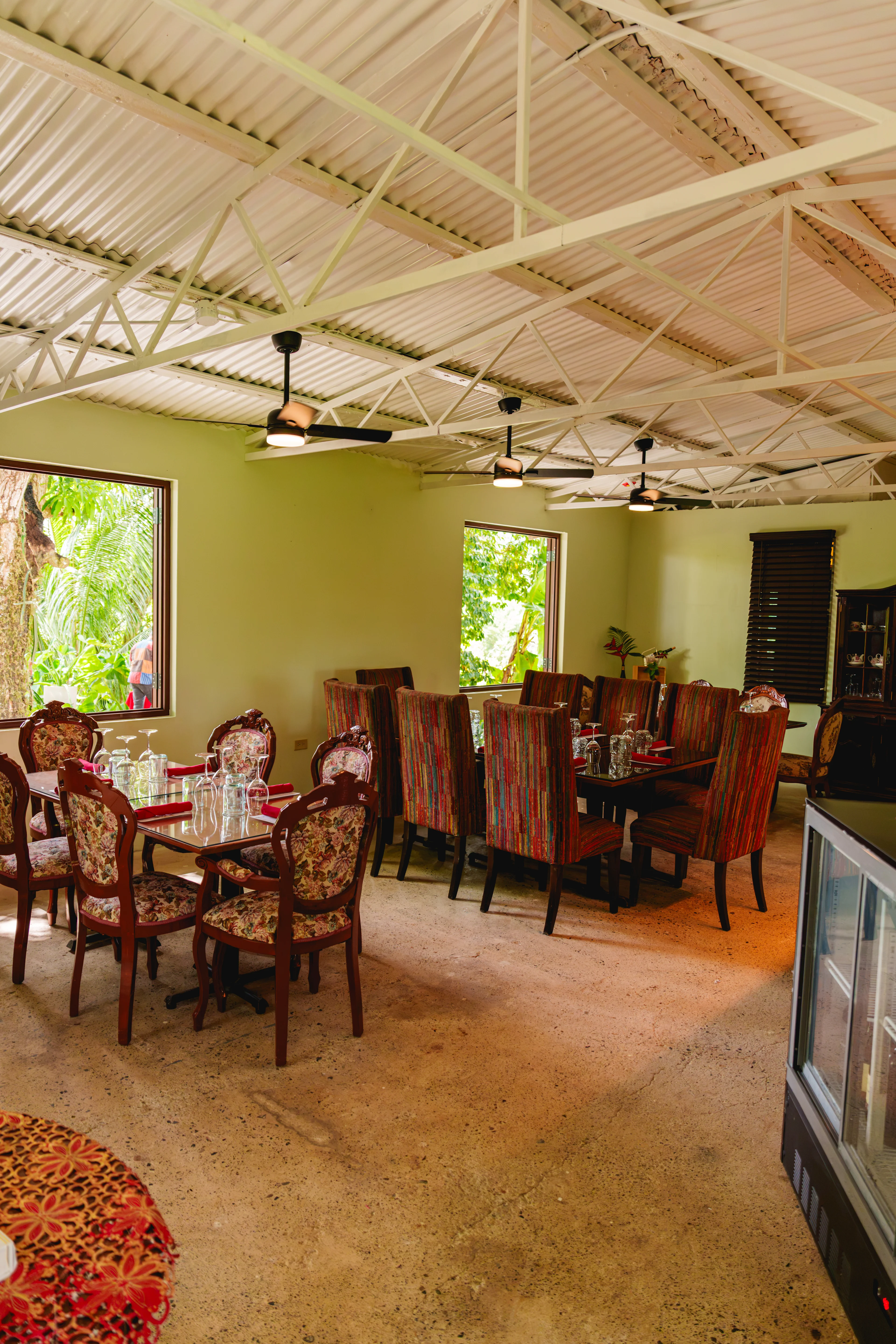 Comedor con paredes verde oliva, techo de zinc con vigas, dos mesas con sillas tapizadas y ventanas con vista a vegetación