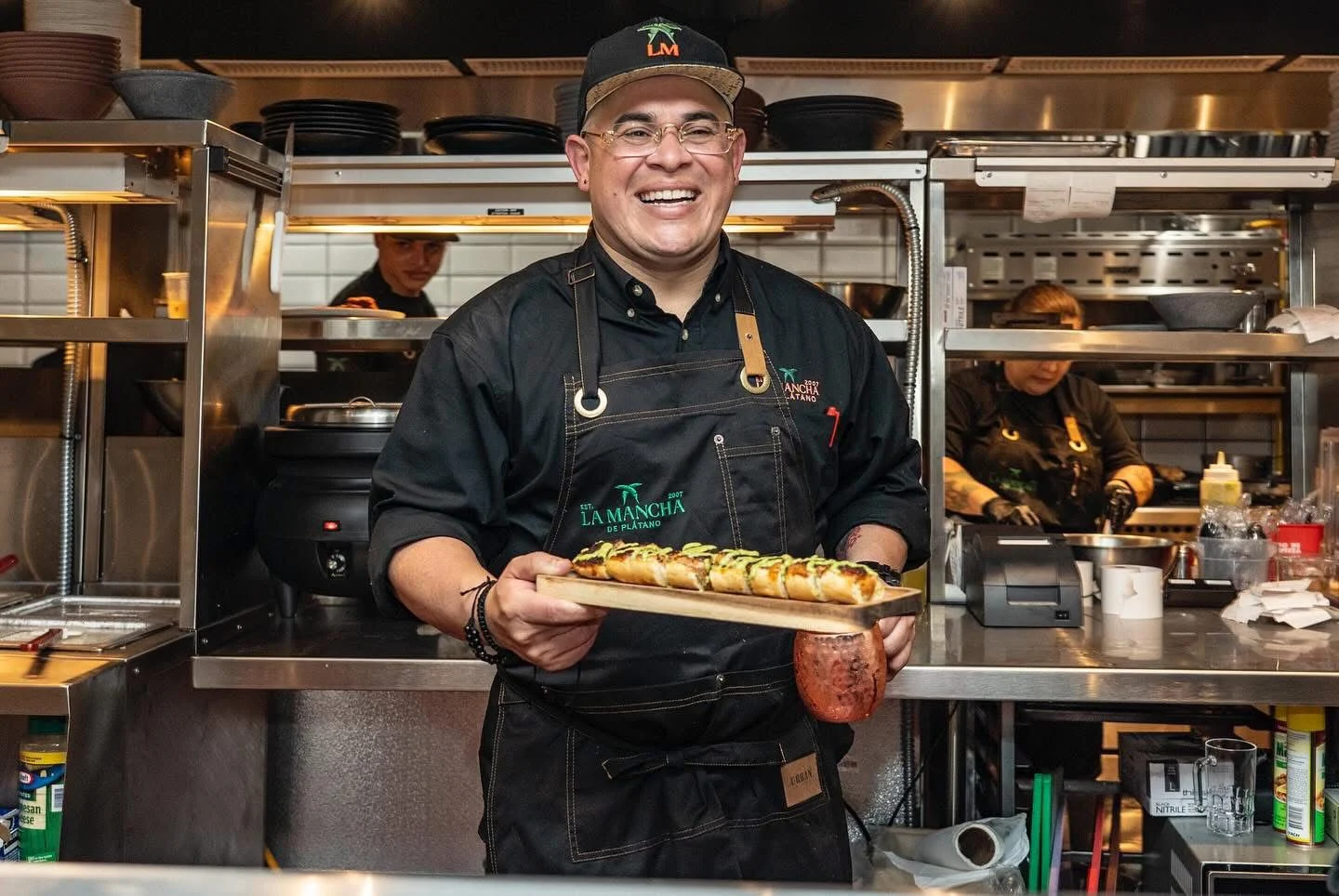 Chef sonriente con gorra y delantal negro sostiene una tabla con plátanos rellenos en la cocina de La Mancha del Plátano
Emanuel Ocasio, chef ejecutivo de La Mancha de Plátano