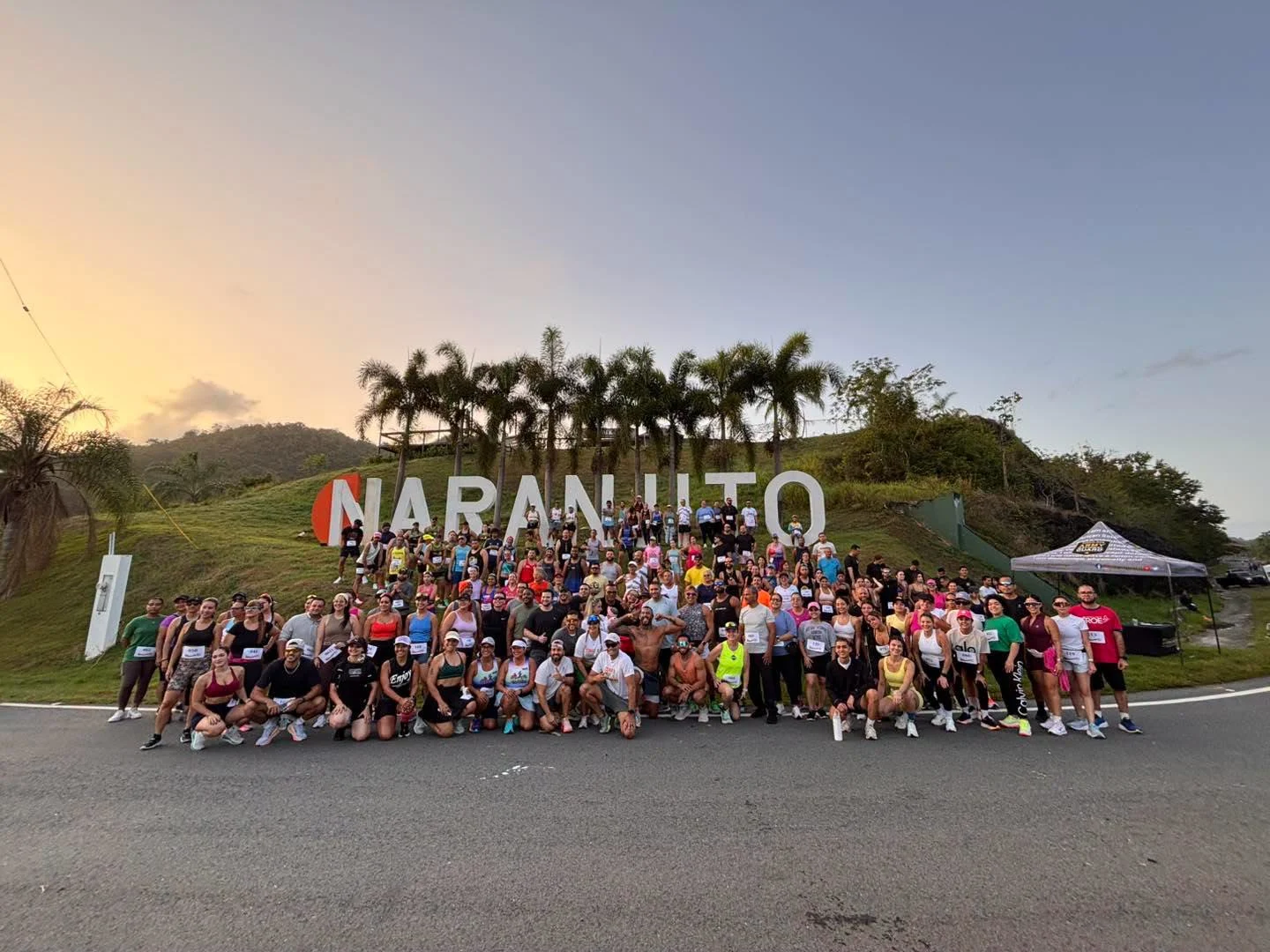 Grupo grande de corredores posa frente al letrero de Naranjito con palmas y montañas verdes al fondo al amanecer
Foto vía Facebook: Municipio Naranjito