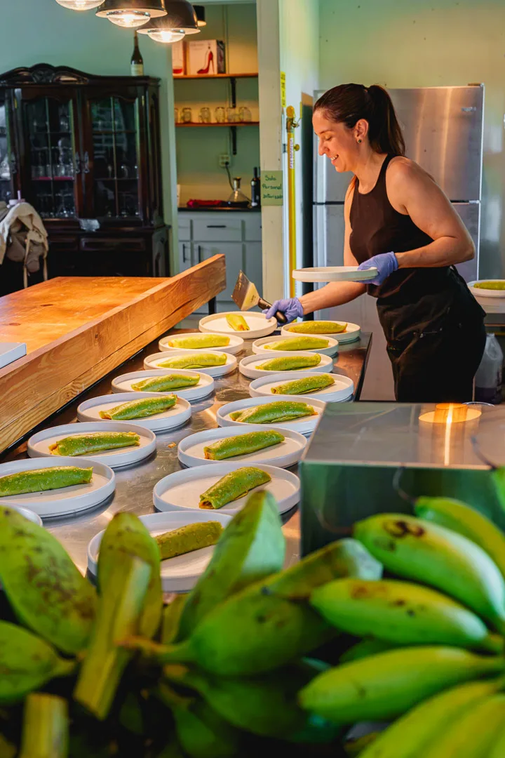 Mujer prepara pionono en cocina con plátanos maduros en primer plano y múltiples platos listos para servir