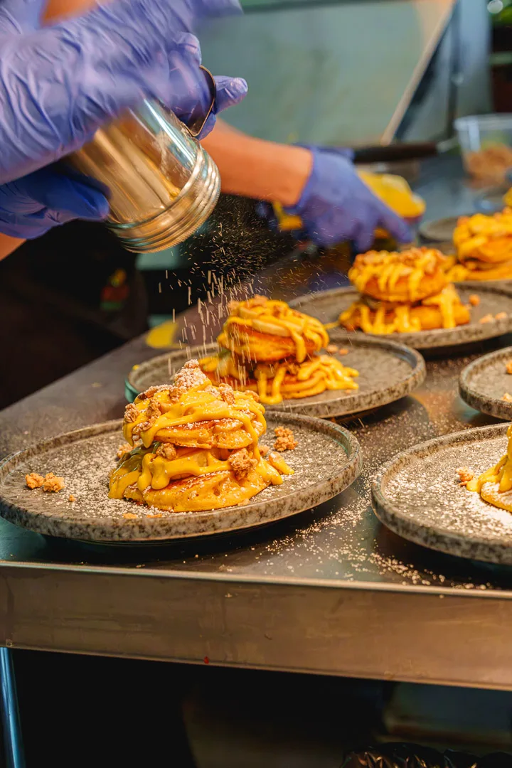 Manos con guantes espolvoreando azúcar sobre platos de waffles con sirope amarillo en una plancha de cocina