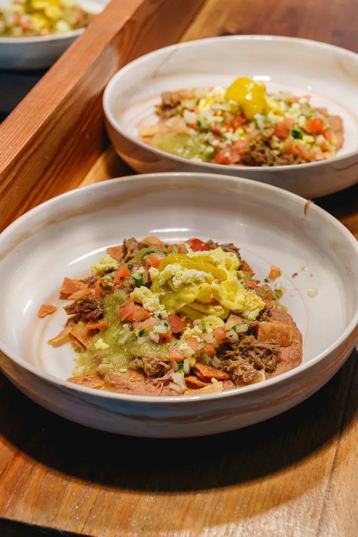 Dos platos hondos con carne mechada, tostones, pico de gallo, salsa verde y plátanos amarillos sobre tabla de madera