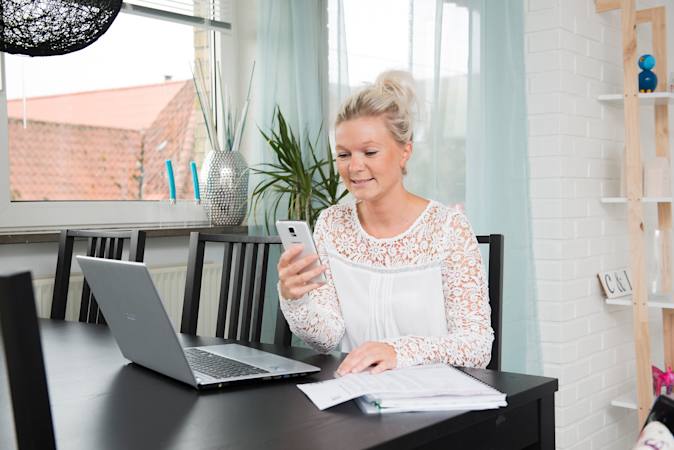 Carina Overgaard sidder ved et bord foran en computer og med en mobiltelefon i hånden