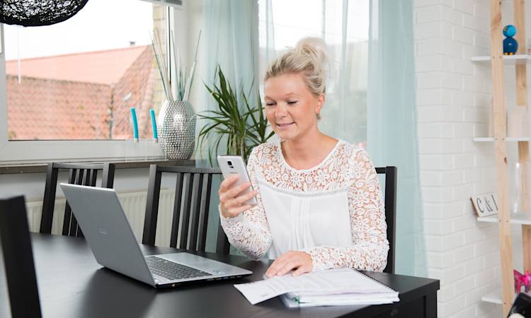 Carina Overgaard sidder ved et bord foran en computer og med en mobiltelefon i hånden