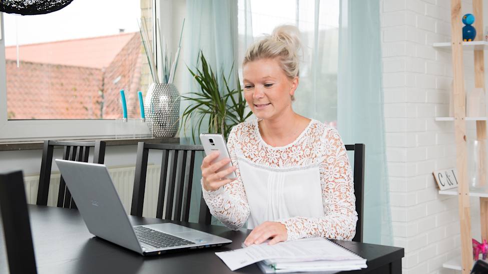 Carina Overgaard sidder ved et bord foran en computer og med en mobiltelefon i hånden