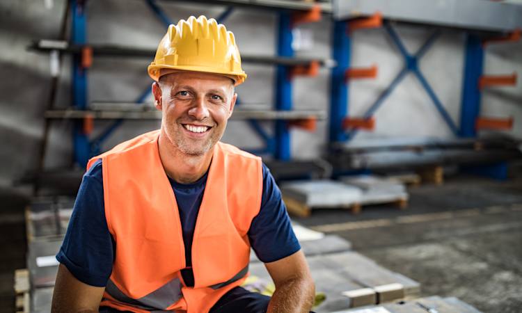 Glad medarbejder sidder på nogle trævare på et lager iført arbejdsvest og hjelm