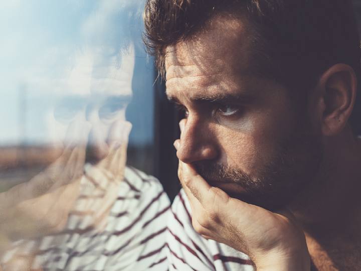 Foto af mand, der med et trist udtryk og den ene hånd op til hovedet kigger ud af vindue