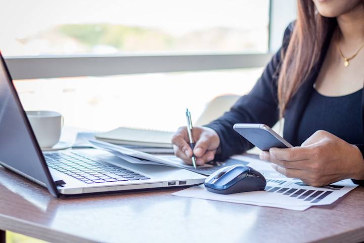 Kvinde ved computer med mobiltelefon og pen i hånden
