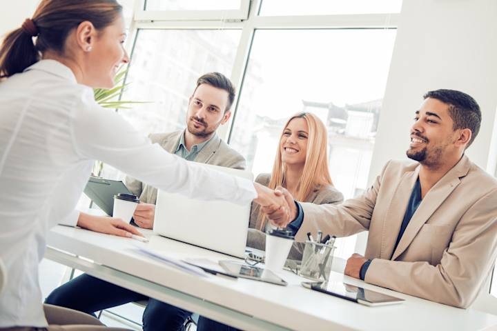 En person sidder foran tre personer ved bord og giver hånd til en af dem