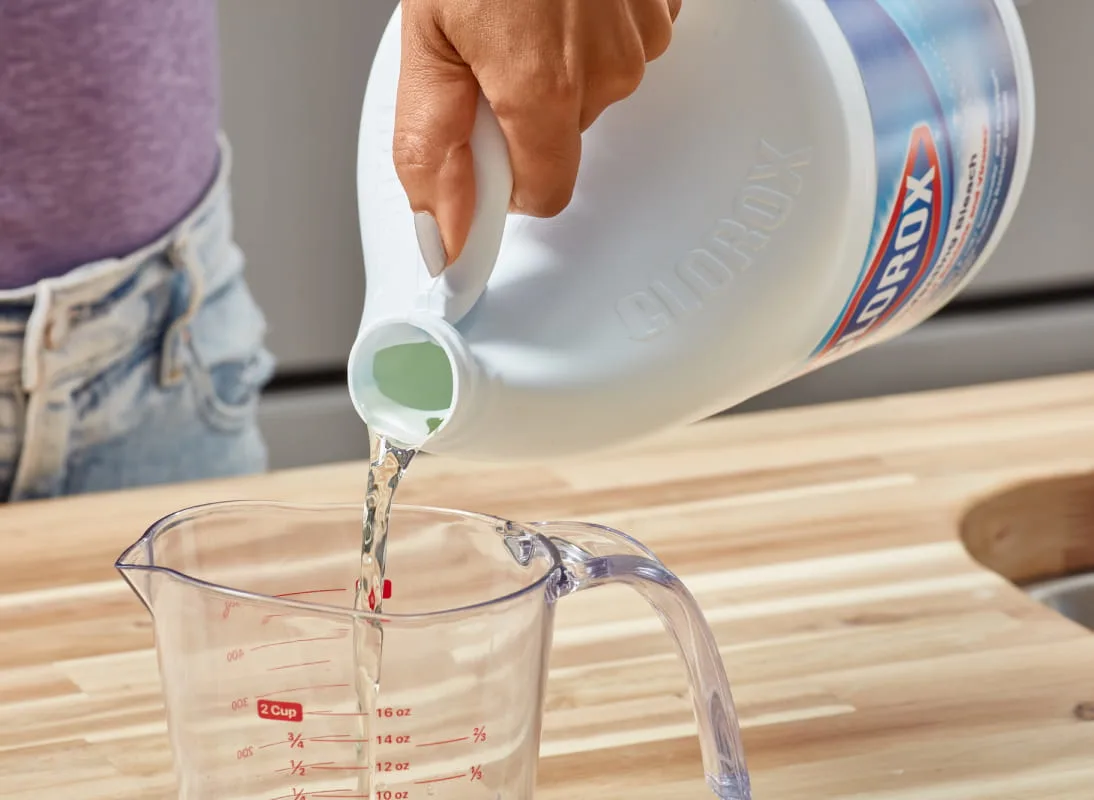 Hand pouring bleach from white Clorox bottle into clear measuring cup on wooden surface