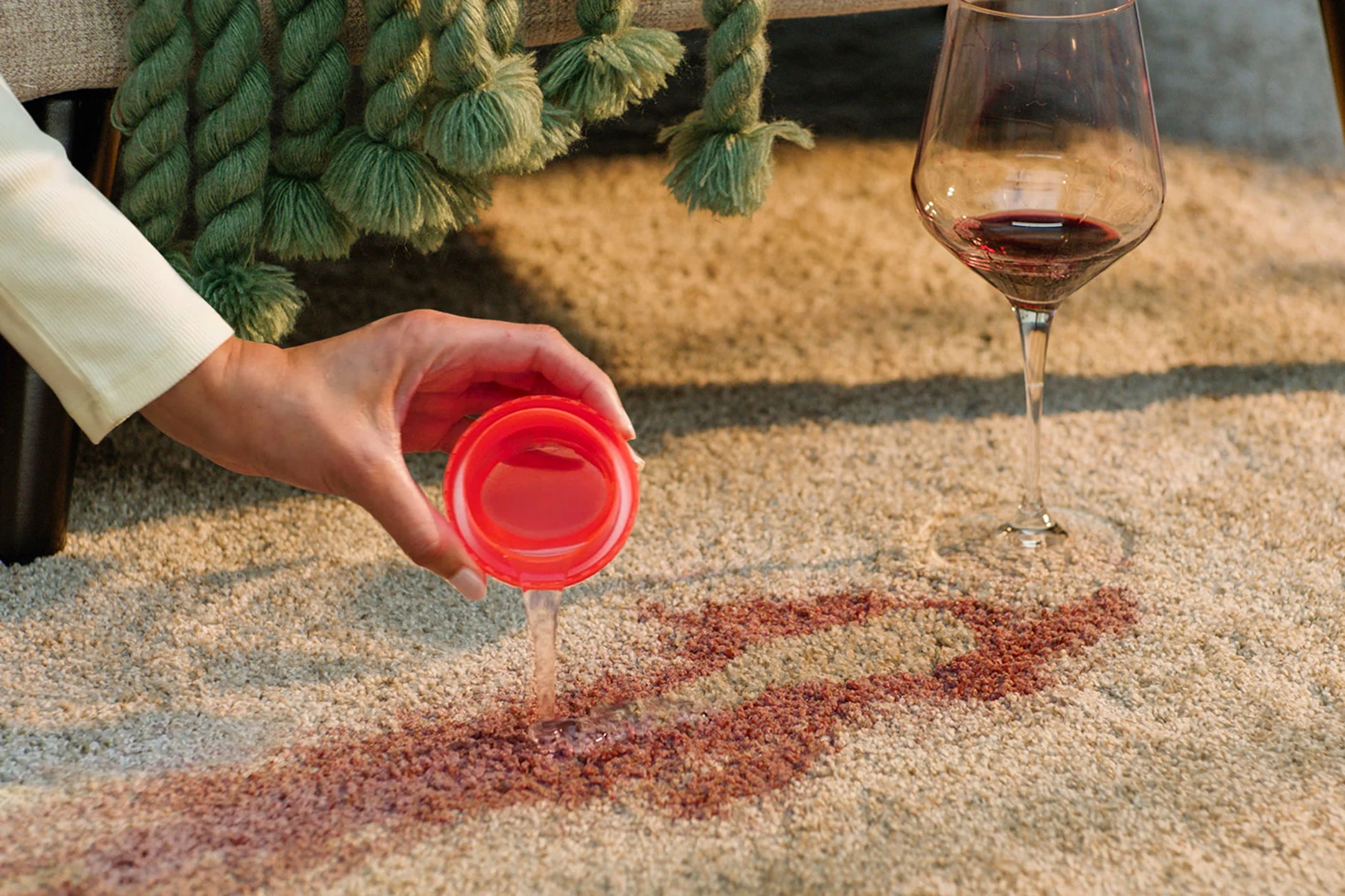 Hand holding red cap over wine stain on carpet, wine glass and green decorative rope nearby.