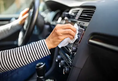 Female wearing long-sleeve striped shirt sitting in front seat, wiping car dashboard with white cloth.