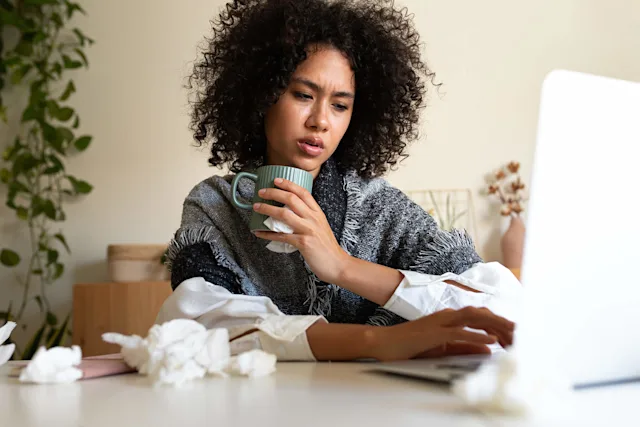 Persona con cabello rizado bebiendo de una taza verde mientras trabaja en una computadora portátil, rodeada de pañuelos arrugados en un escritorio.