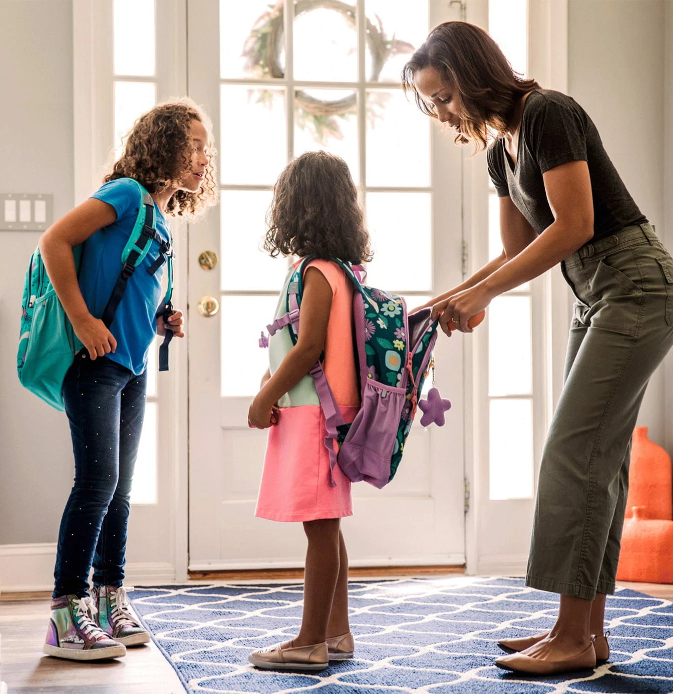 Mother helping two daughters with colorful backpacks prepare for school by a bright front door