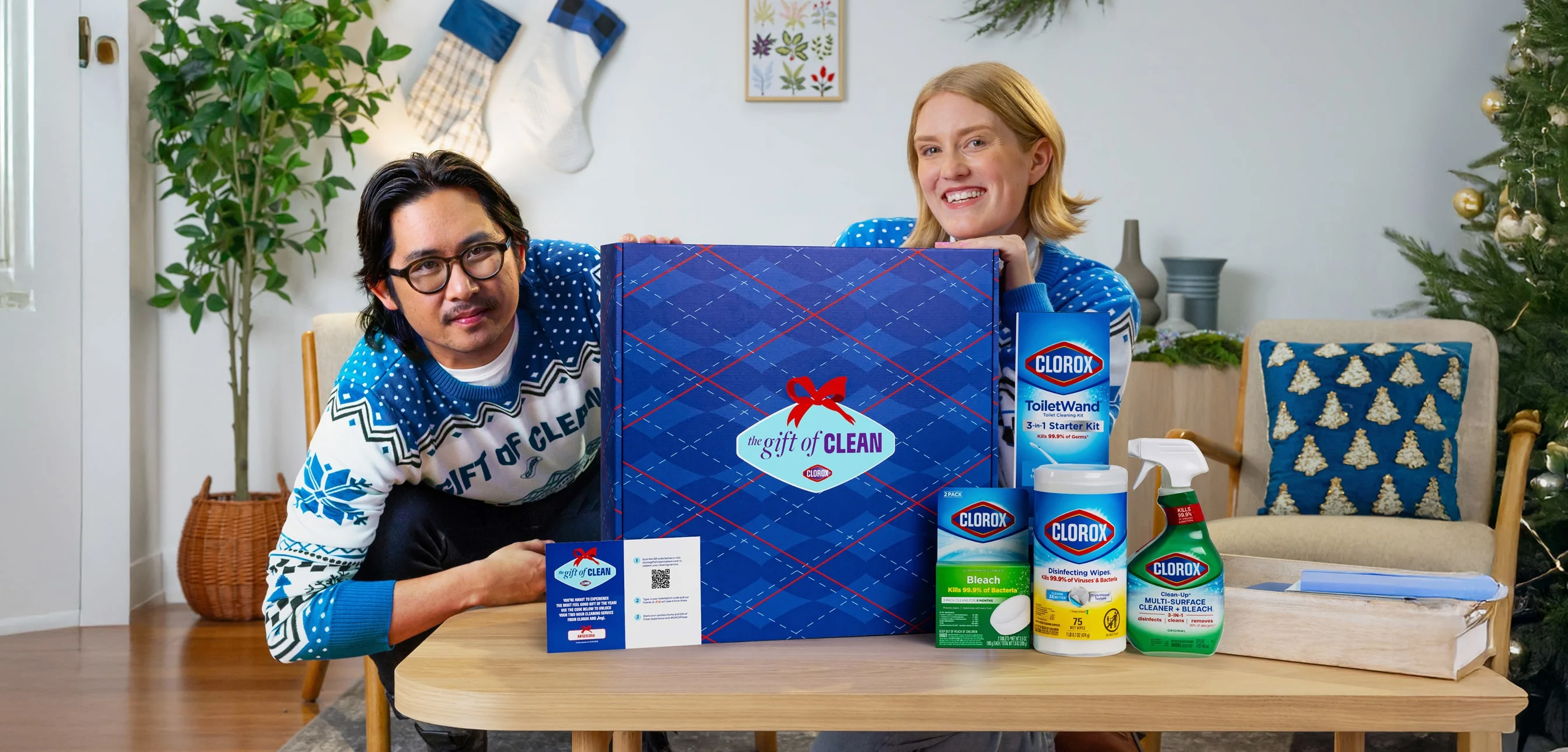 Two people in Gift of Clean holiday sweaters posing with the Gift of Clean box in a living room
