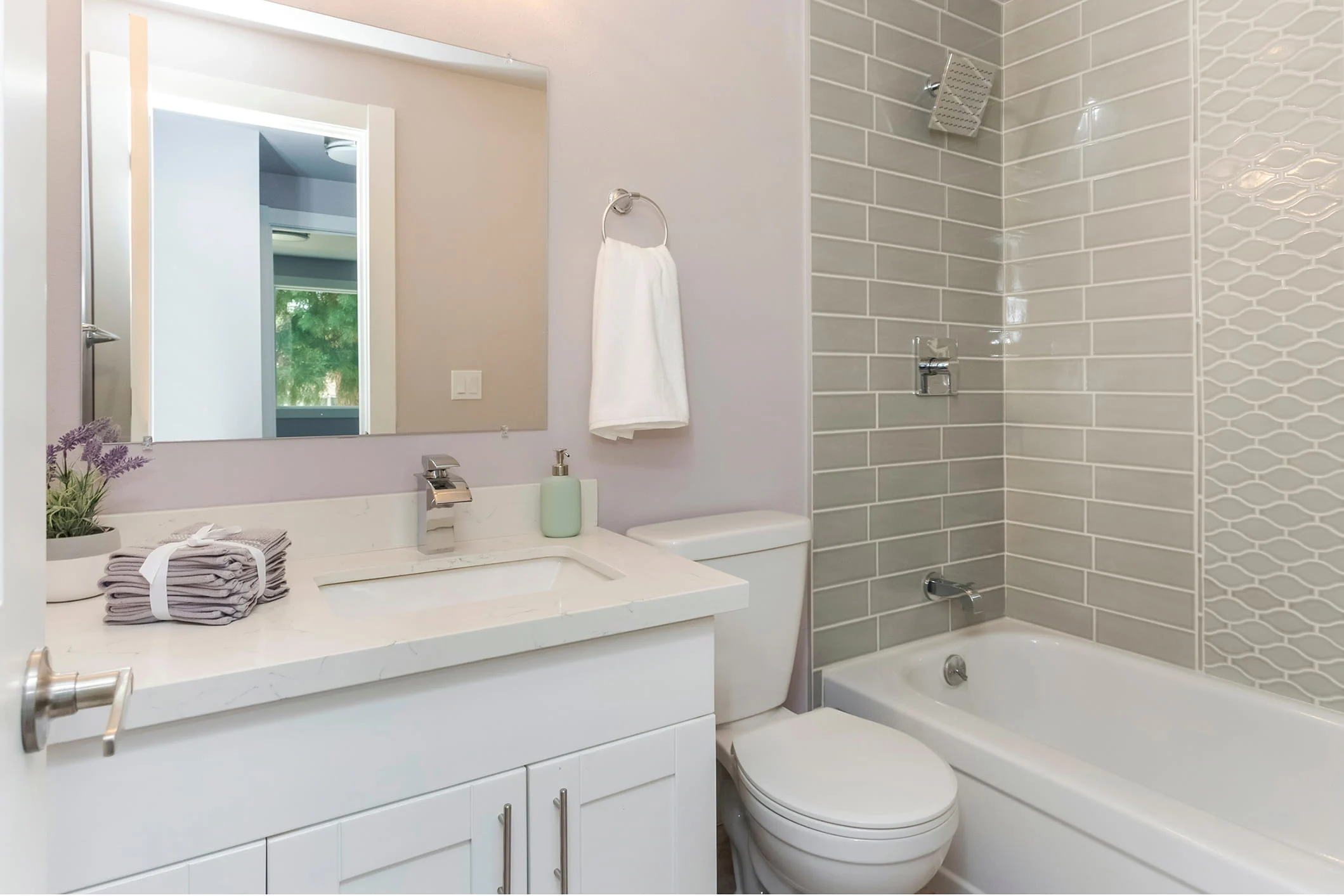 Baño moderno con tocador blanco, ducha con azulejos beige tipo metro, inodoro blanco y espejo sobre el lavabo con toallas dobladas.