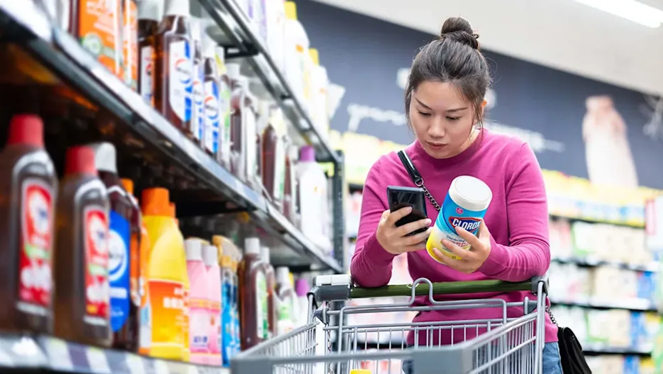 Woman leaning on shopping cart in store, with phone and cleaning product in hand, reviewing details.