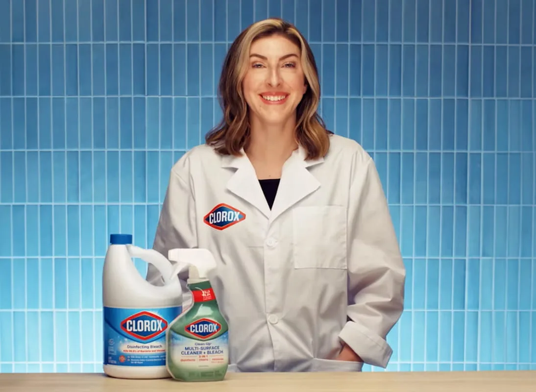 A woman in a lab coat with a bottle of disinfecting bleach and a spray bottle of Clorox Clean-Up on the table