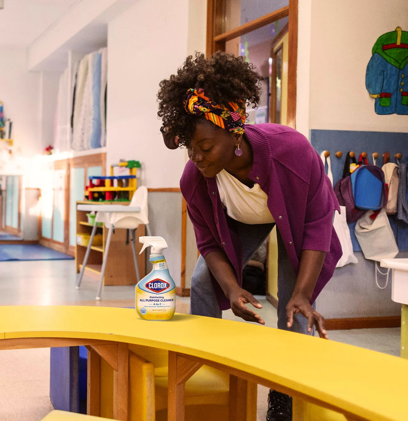 Woman in purple cardigan and headband cleaning a yellow table with Clorox spray in a classroom.