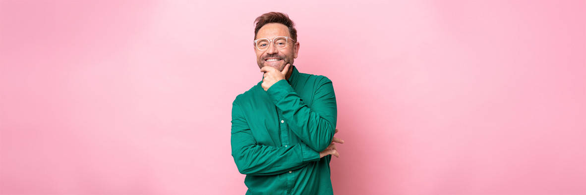 A bearded man in a green shirt smiles broadly against a pink studio background.