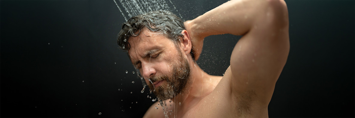 A bearded man lathers shampoo through his hair in the shower with eyes closed.
