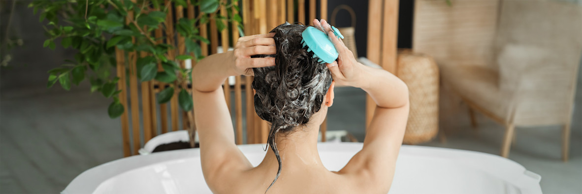 A woman washes her hair outdoors against a teal background, lather visible.
