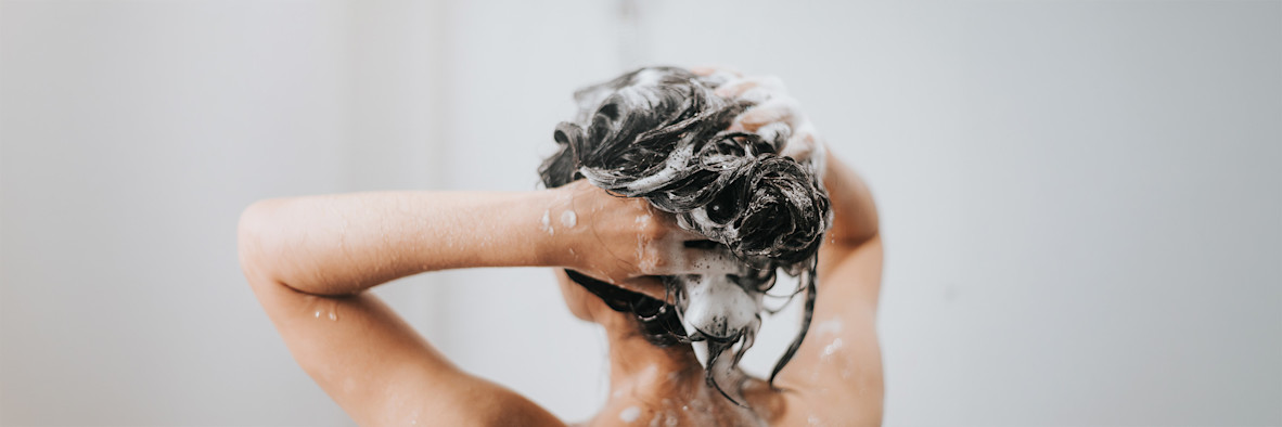 A woman massages shampoo through her hair, eyes closed, with white lather visible.