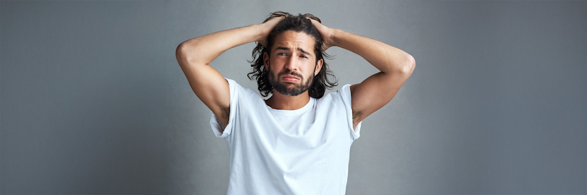A bearded man in a white t-shirt clasps his hands behind his head looking upward.