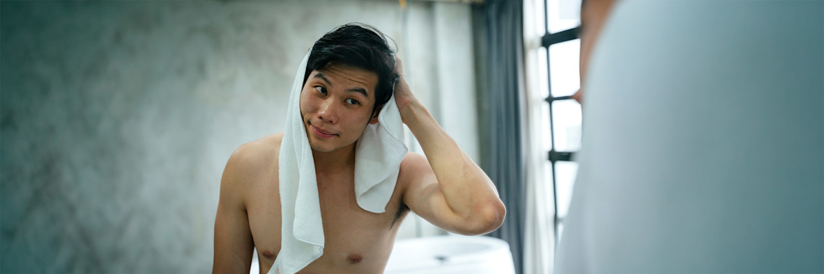 A man wraps his hair in a white towel after washing, reflected in a bathroom mirror.