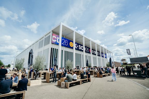 Im Hintergrund ist der Veranstaltungsort der SCD, das Confex, vor einem hellblauen Himmel mit weißen Wolken zu sehen. Im Vordergrund sitzen Menschen an Tischen auf Holzbänken und essen zu Mittag. Die Atmosphäre wirkt entspannt und gemütlich.