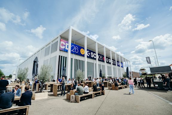 Confex SCD 2026 Im Hintergrund ist der Veranstaltungsort der SCD, das Confex, vor einem hellblauen Himmel mit weißen Wolken zu sehen. Im Vordergrund sitzen Menschen an Tischen auf Holzbänken und essen zu Mittag. Die Atmosphäre wirkt entspannt und gemütlich.