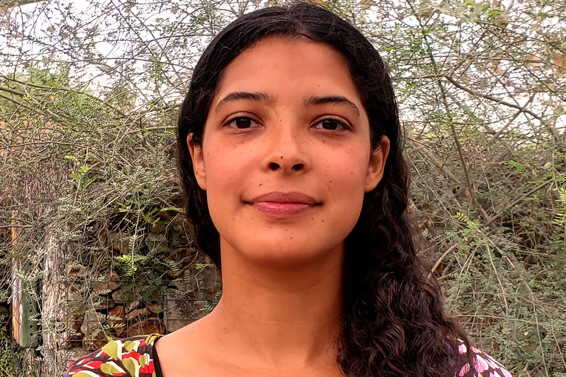 A woman poses against a background of tall green plants.